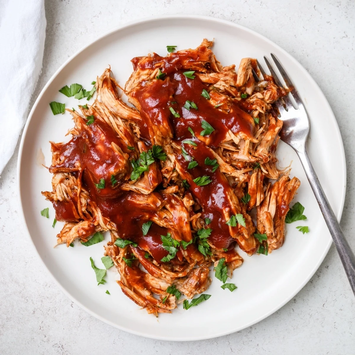 Tender shredded chicken pieces smothered in sticky sweet BBQ sauce on white serving plate
